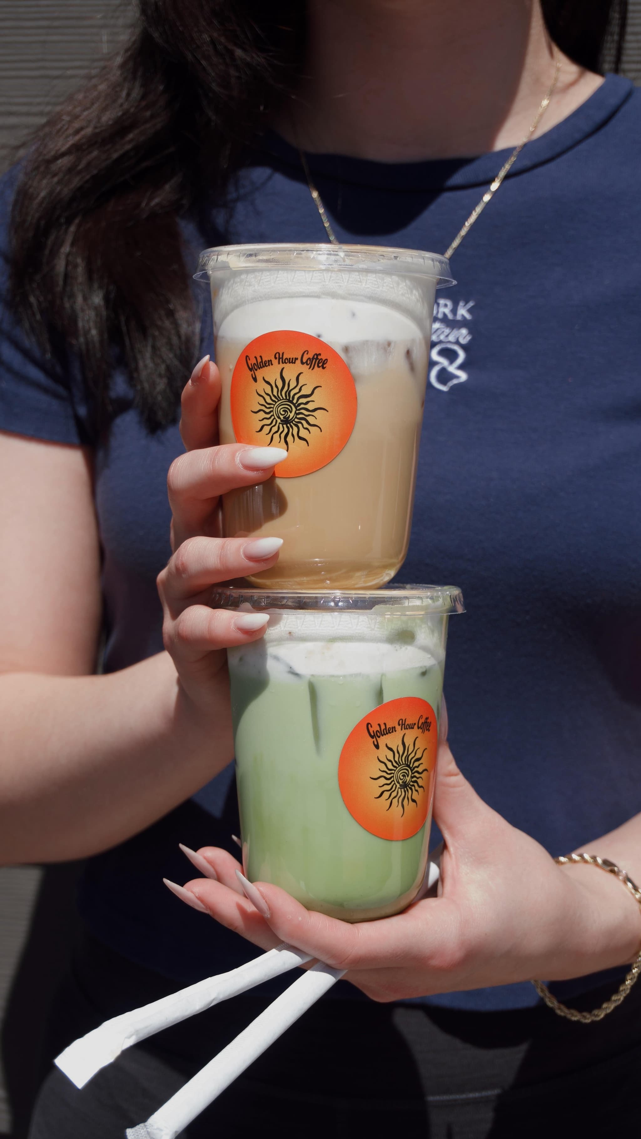 Two stacked Golden Hour cups held up in the sun, an iced latte on top and an iced matcha below, each wearing the orange sun sticker.