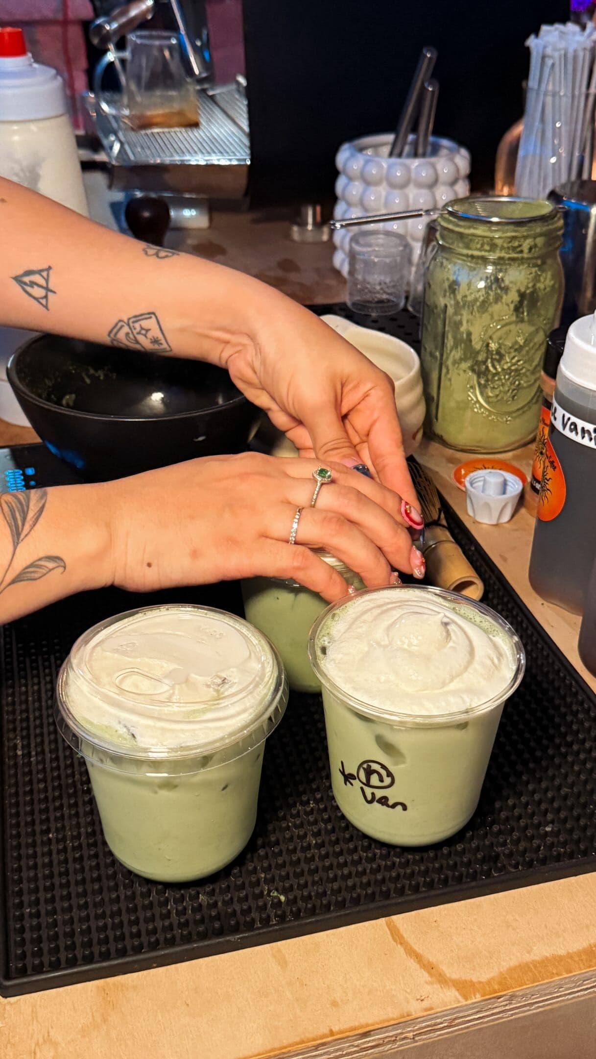 A barista's tattooed, ringed hands topping two iced matcha lattes with cold foam at the cart.