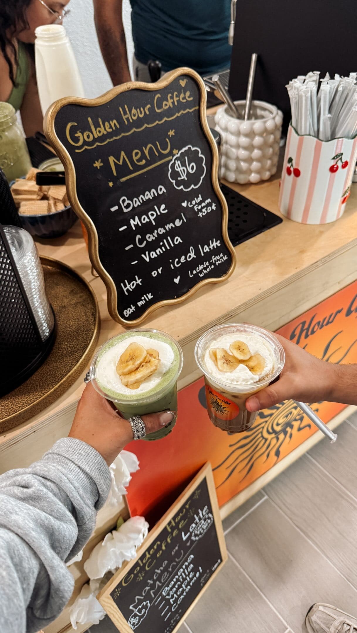 A banana matcha and a banana latte cheersed over the counter in front of the handwritten Golden Hour Coffee menu board and the orange sun cart banner.